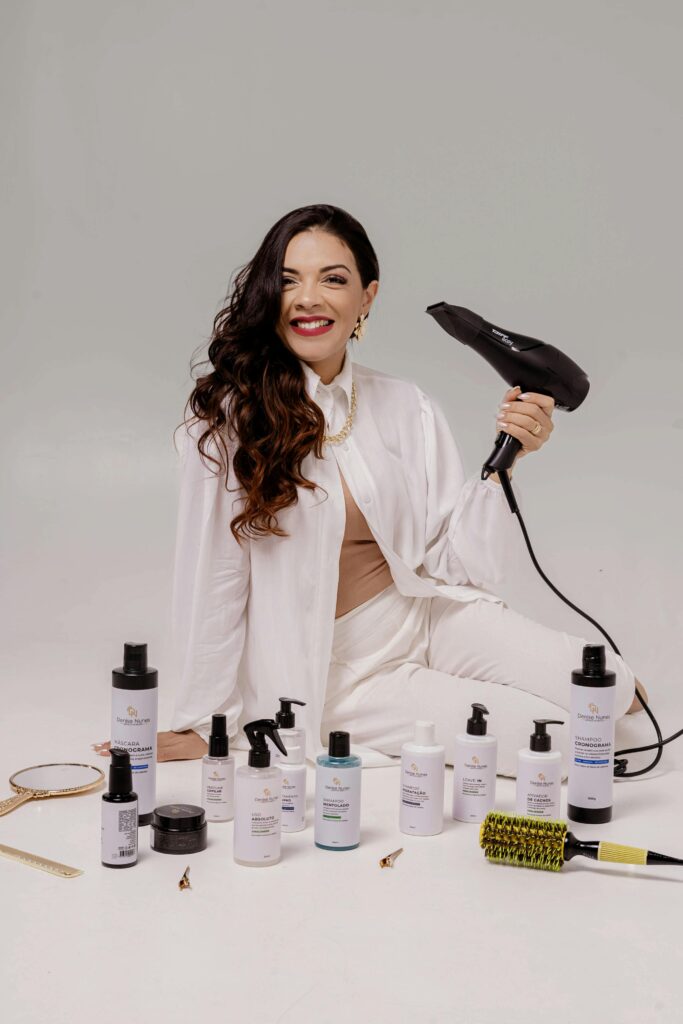 Smiling woman holding a hairdryer, surrounded by various hair care products.